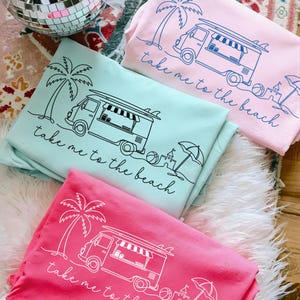 May include: Three sweatshirts in shades of pink and teal, each featuring a beach-themed graphic. The design includes a palm tree, a van with a surfboard, and the phrase "take me to the beach."