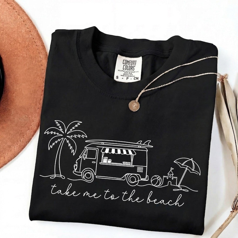 May include: Black Comfort Colors t-shirt with a white graphic of a food truck, palm tree, beach ball, sandcastle, and umbrella. The text "take me to the beach" is below the graphic. A gold necklace with a pendant is on the shirt.