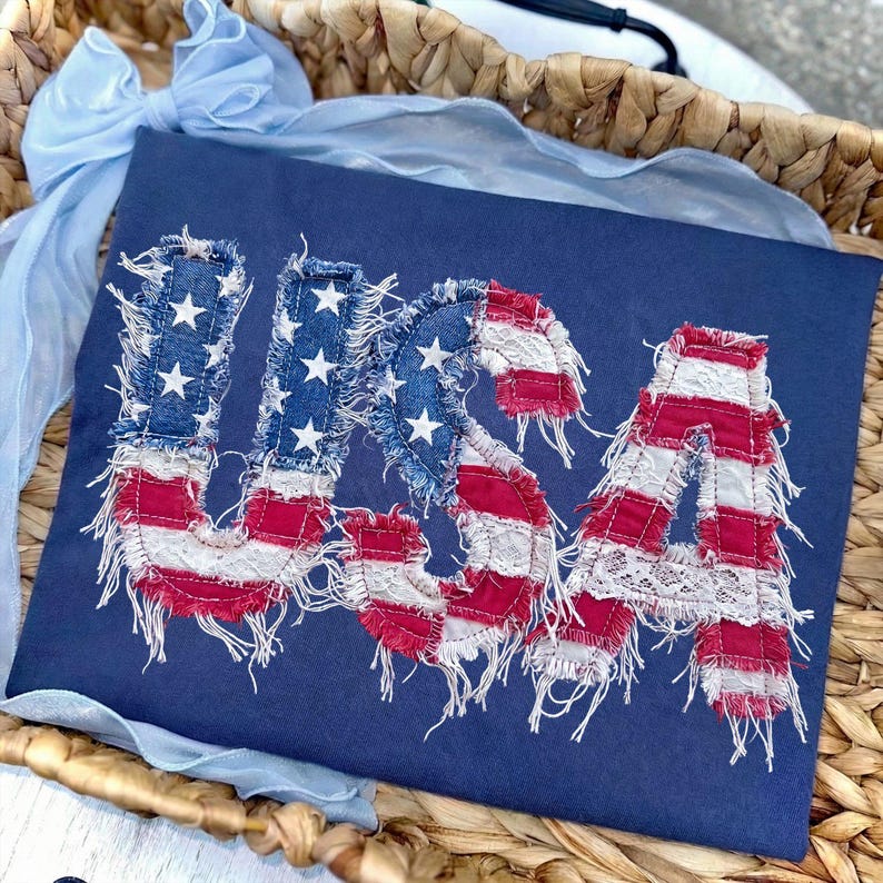 May include: Navy blue t-shirt featuring the word "USA" in a distressed American flag design. The letters are made of denim and red and white striped fabric, with white stars on the "U" and "S". A light blue ribbon is in the background.