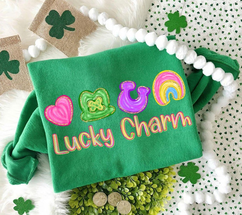 May include: A green sweatshirt with a "Lucky Charm" design featuring a pink heart, a green shamrock, a purple horseshoe, and a rainbow. The design has a yellow and pink outline. The sweatshirt is surrounded by decorative elements.