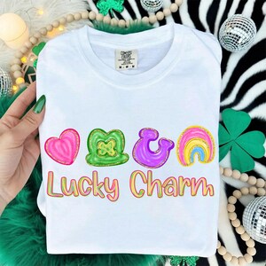May include: White crew neck t-shirt with a colorful "Lucky Charm" design. The design includes a pink heart, green shamrock, purple horseshoe, and rainbow. The text is in a colorful, glittery font.