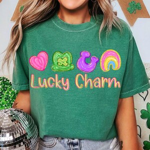 May include: Green t-shirt with the words "Lucky Charm" in a colorful, bubble-letter font. The shirt features four colorful graphic designs: a pink heart, a green shamrock, a purple horseshoe, and a rainbow.
