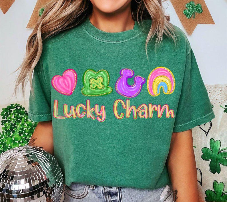 May include: Green t-shirt with the words "Lucky Charm" in a colorful, bubble-letter font. The shirt features four colorful graphic designs: a pink heart, a green shamrock, a purple horseshoe, and a rainbow.