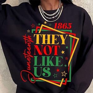 Peut inclure: Sweat-shirt noir avec l'inscription "Juneteenth" en rouge. Le motif comprend les chiffres "1865" et la phrase "THEY NOT LIKE US" en jaune, rouge et vert, avec des étoiles.