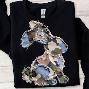 Può includere: Felpa nera con un design a coniglio patchwork. Il coniglio è realizzato in tessuto mimetico e denim con bordi sfilacciati. La felpa ha un girocollo e maniche lunghe.