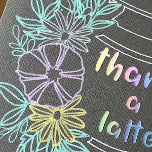 Peut inclure: Carte grise foncée avec un motif floral bleu clair, jaune et violet. Les mots "thank a latte" sont écrits en lettres irisées. La carte a une surface texturée.
