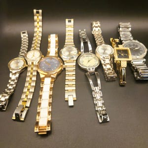 May include: A selection of women's wristwatches with silver, gold, and white straps. The watches feature round and square faces with diverse dial colours. Some watches have decorative features.