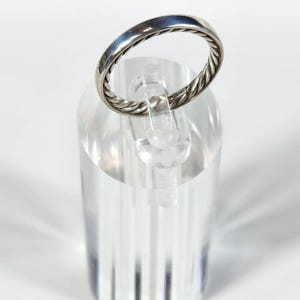May include: A silver ring with a twisted rope design around the band. The ring is displayed on a clear acrylic stand against a white background. The ring is a simple, elegant piece of jewelry.