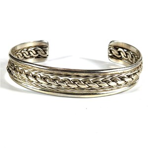 May include: Silver-toned cuff bracelet with a braided design. The bracelet features a thick, open design with a series of interwoven links. The metal has a polished finish, reflecting light. The bracelet is a jewelry item.