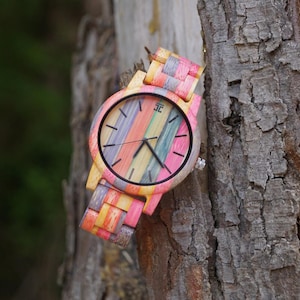 Puede incluir: Reloj de pulsera de madera multicolor con esfera y correa a rayas. El reloj tiene una esfera negra con manecillas negras y una corona plateada. El reloj está hecho de madera y está sujeto a una correa de madera.