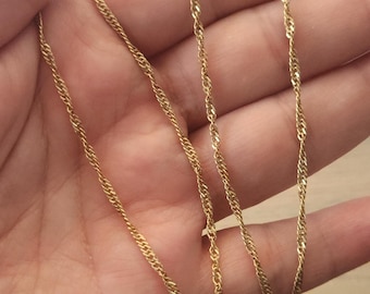 Puede incluir: Un collar de cadena de cuerda retorcida de color dorado. La cadena se muestra sobre un fondo claro, mostrando su intrincado diseño y brillo metálico. El collar es una pieza de joyería delicada.