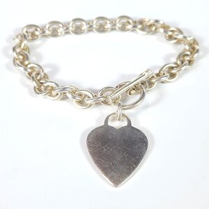 May include: A silver-toned chain link bracelet featuring a toggle clasp closure and a single heart-shaped pendant charm. The metal surface of the heart charm displays visible fine scratches and a smooth, reflective finish throughout the piece.