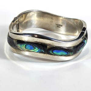 May include: A silver hinged bracelet with iridescent abalone shell inlays. The bracelet has a curved design and is marked "MEXICO 925". The abalone shell displays shades of blue, green, and black.