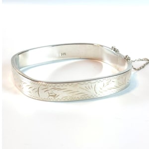 May include: A silver-colored hinged bangle bracelet with a rectangular shape and rounded corners. The exterior surface features an engraved leaf-like pattern along the center. The interior is stamped with the number 925 and includes a safety chain.
