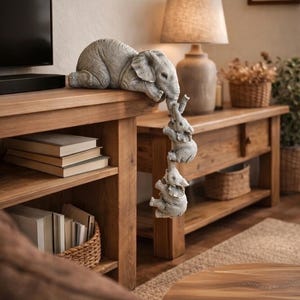 May include: A gray elephant family sculpture. The largest elephant rests on a wooden surface, with three smaller elephants hanging below, each holding the tail of the one above. The sculpture is a decorative accent for a home.