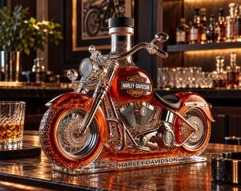 May include: A clear glass decanter shaped like a motorcycle filled with amber liquid. The fuel tank features the Harley-Davidson Motor Company logo in orange and black. The base is inscribed with the text Harley-Davidson in black lettering.