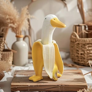 Pode incluir: Uma figura decorativa extravagante de um pato com corpo de banana. O pato tem cabeça e pescoço brancos com bico e pés amarelos. A banana é amarela com detalhes brancos, em cima de uma tábua de madeira.