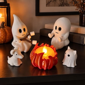 Op de afbeelding: Een set witte spookbeeldjes, één met een stokje met twee marshmallows, een andere zittend, een kleine witte hond, een kleine witte kat en een centrale oranje en rode vlamvormige lamp met een gloeiende gele LED-vlam erin.
