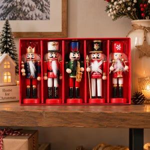 Può includere: Un set di cinque statuette schiaccianoci in legno presentate in una scatola rossa. Le figure includono un re con corona, un soldato in rosso, un musicista con sassofono, un soldato con bastone e un tamburino, tutti con barbe bianche e uniformi.