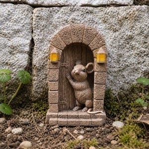 Puede incluir: Un adorno de jardín caprichoso que presenta un ratón marrón detallado asomándose por una puerta arqueada en miniatura. La puerta de madera tiene dos pequeñas linternas a cada lado. La escena está ambientada contra una pared de piedra y vegetación.