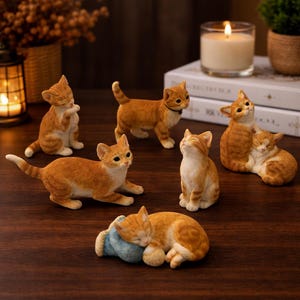 Puede incluir: Una colección de figuras de gatos de color naranja y blanco en varias poses, incluyendo de pie, sentados y acostados. Un gato está acurrucado durmiendo sobre una almohada azul. Las figuras están dispuestas sobre una superficie de madera oscura.