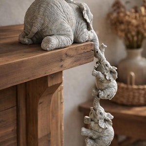 May include: A gray elephant sculpture with three smaller elephants hanging from its trunk. The sculpture is resting on a wooden surface. The elephants are detailed with textured skin and are a light gray color.