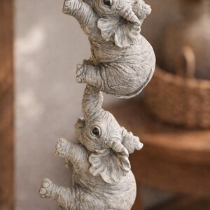 May include: Three gray elephant figurines are stacked, with each one holding onto the other. The elephants have detailed features, including wrinkles and large ears. The figurines appear to be made of stone or a similar material.