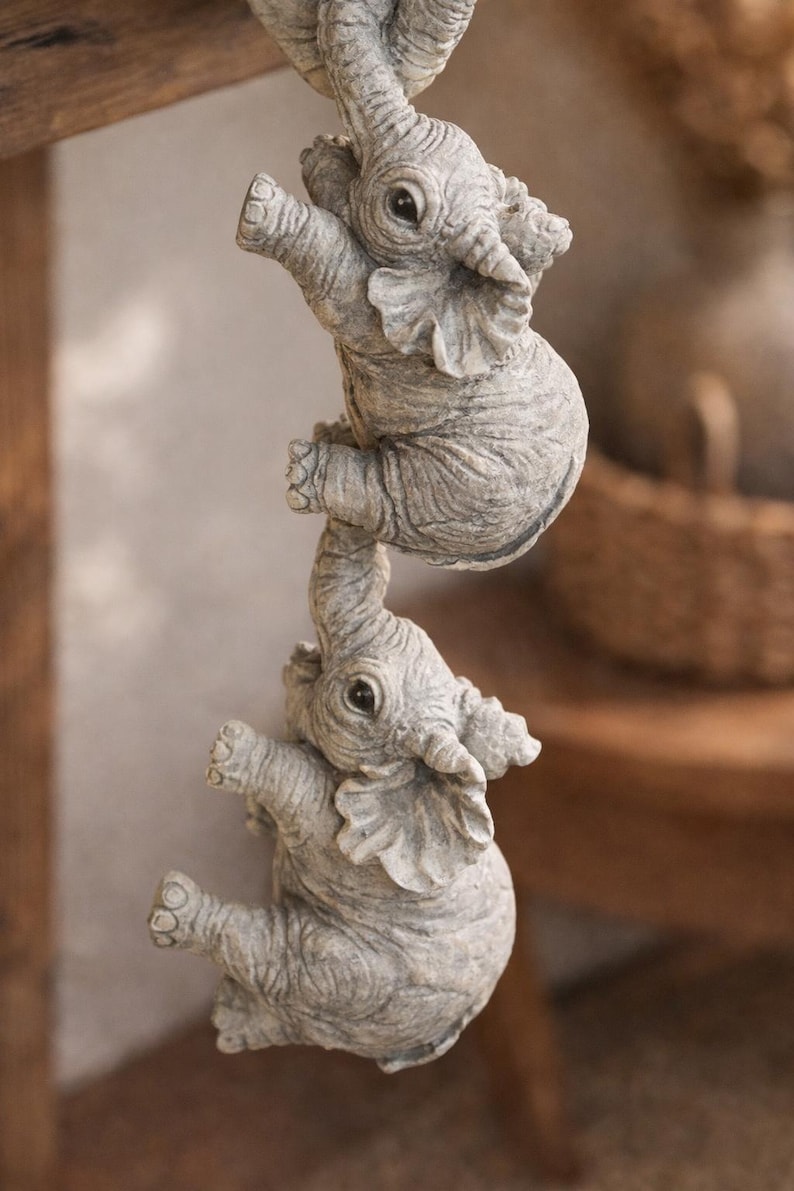 May include: Three gray elephant figurines are stacked, with each one holding onto the other. The elephants have detailed features, including wrinkles and large ears. The figurines appear to be made of stone or a similar material.