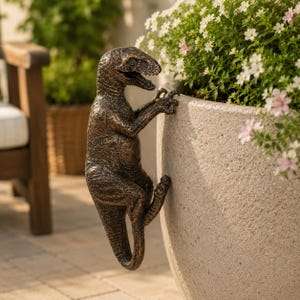 Peut inclure: Sculpture de dinosaure de couleur bronze grimpant sur un pot de fleurs clair. La sculpture présente des détails précis et est positionnée sur le côté du pot, ses griffes agrippant le bord. Des fleurs blanches et roses sont visibles dans le pot.