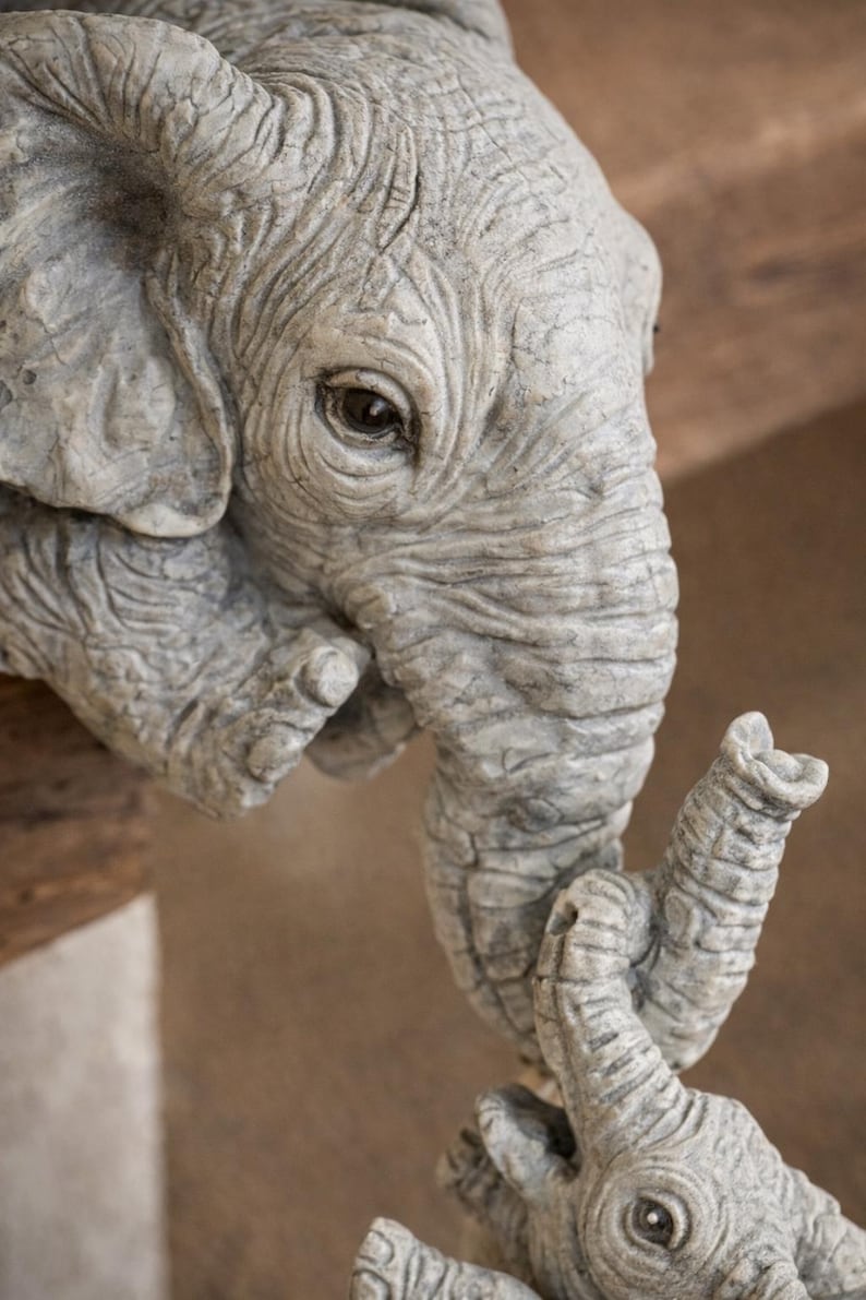 May include: A detailed sculpture of an elephant head and trunk, with a smaller elephant calf. The sculpture is a light gray color, with visible wrinkles and textures. The trunk of the larger elephant is touching the trunk of the smaller elephant.