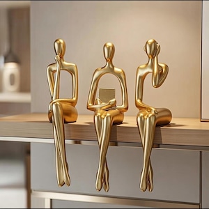 Pode incluir: Três esculturas abstratas douradas de figuras sentadas. Cada figura tem uma pose única: uma com a mão no queixo, outra segurando um pequeno quadrado e a terceira cobrindo a orelha. As esculturas são exibidas em uma prateleira de madeira.