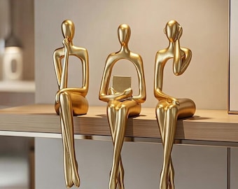 May include: Three gold-colored abstract sculptures of seated figures. Each figure has a unique pose: one with a hand on the chin, another holding a small square, and the third covering their ear. The sculptures are displayed on a wooden shelf.