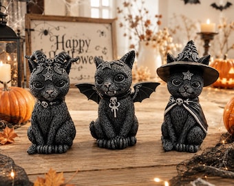 Peut inclure: Trois figurines de chats noirs aux textures détaillées. Le chat de gauche a des cornes et un pentagramme. Le chat central possède des ailes de chauve-souris et un pendentif crâne. Le chat de droite porte un chapeau de sorcière et une cape.