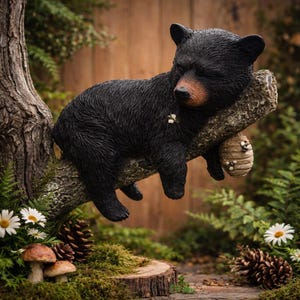 May include: A black bear cub figurine resting on a tree branch. The bear has a brown snout and is positioned above a small beehive. The scene includes daisies, pine cones, and mushrooms, creating a woodland theme.