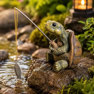 Op de afbeelding: Gedetailleerd beeldje van een schildpad die aan het vissen is. De schildpad is groen en bruin en houdt een hengel vast met een zilveren vislokmiddel. Het beeldje staat op een rots, met een beekje en groen op de achtergrond.