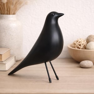 Puede incluir: Una figura decorativa de pájaro negro mate con un diseño elegante y minimalista. El pájaro tiene un cuerpo redondeado, un pico puntiagudo y se apoya sobre dos finas patas metálicas. Un acento moderno para la decoración del hogar.