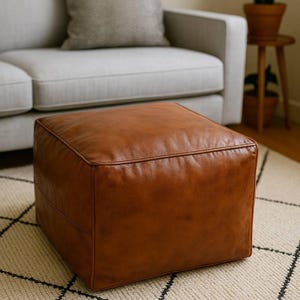 May include: A square ottoman upholstered in smooth, cognac-colored leather. The piece features clean, straight edges, a flat top surface, and visible piping along the seams, creating a structured, boxy silhouette with a uniform, warm brown finish.