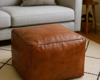 May include: A square ottoman upholstered in smooth, cognac-colored leather. The piece features clean, straight edges, visible piping along the top perimeter, and a structured, boxy silhouette with a uniform, solid-toned finish across all visible sides.