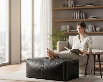 May include: A black leather ottoman with white stitching. A person is sitting on a light-coloured sofa, reading a book, with their feet resting on the ottoman. A small side table and a window are also visible in the living room.