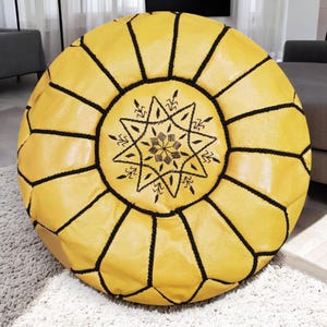 May include: A round, yellow leather pouf with black stitching and a central starburst design. The pouf is a decorative piece of furniture, likely used as a footrest or extra seating. The design features geometric patterns.