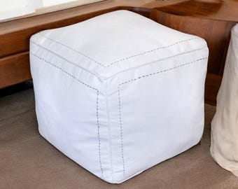 Op de afbeelding: Een witte, kubusvormige poef met gestikte details. De naden zijn geaccentueerd met een stippellijnpatroon in een contrasterende grijze kleur. De poef staat op een bruin tapijt.