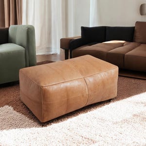 May include: A brown leather ottoman with a rectangular shape and a slightly curved top. The ottoman is sitting on a white rug.