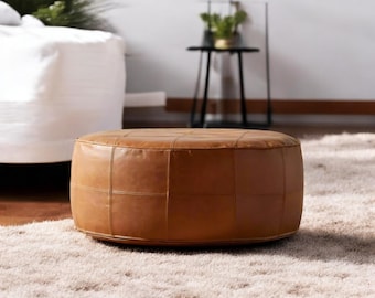 Può includere: Un pouf rotondo in pelle marrone con un design cucito a griglia. Il pouf è posto su un tappeto chiaro e peloso. La stanza ha una palette di colori neutri, con un divano bianco e un tavolino nero sullo sfondo.