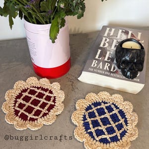 Peut inclure: Deux dessous de verre faits à la main, un bordeaux et un bleu marine, ressemblant à des tartes miniatures. Chaque dessous de verre a un bord festonné beige et un motif en treillis. L'image comprend également un livre, un bougeoir en forme de crâne et un vase de fleurs.