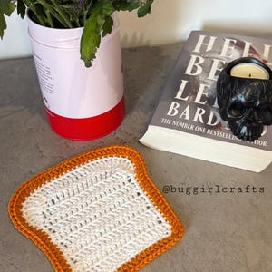 May include: A crochet coaster shaped like a slice of toast, with a white centre and an orange border. The image also shows a book, a skull-shaped candle holder, and a pink and red container with flowers.