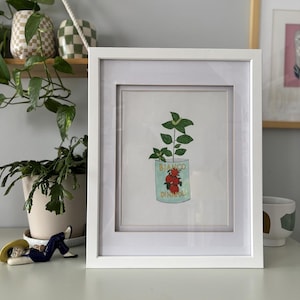 May include: A framed art print featuring a watercolor illustration of a tomato can with a plant growing out of it. The can is light blue with the words "BIANCO DINATOLI" and a cluster of red tomatoes. The frame is white.