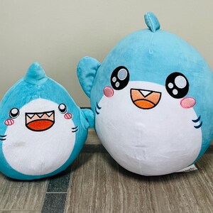 May include: Two plush shark toys. The sharks are light blue with white bellies, black eyes, and smiling mouths. The larger shark has a small yellow fish attached to its fin. The sharks are sitting on a wooden surface.