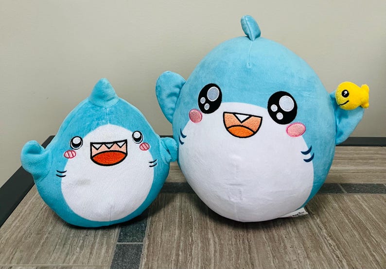 May include: Two plush shark toys. The sharks are light blue with white bellies, black eyes, and smiling mouths. The larger shark has a small yellow fish attached to its fin. The sharks are sitting on a wooden surface.