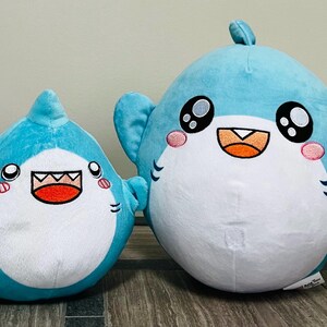 May include: Two plush shark toys in shades of blue and white. The sharks have large, expressive eyes, pink cheeks, and open mouths. One shark has a small yellow fish attached. The toys are designed with a cartoonish aesthetic.