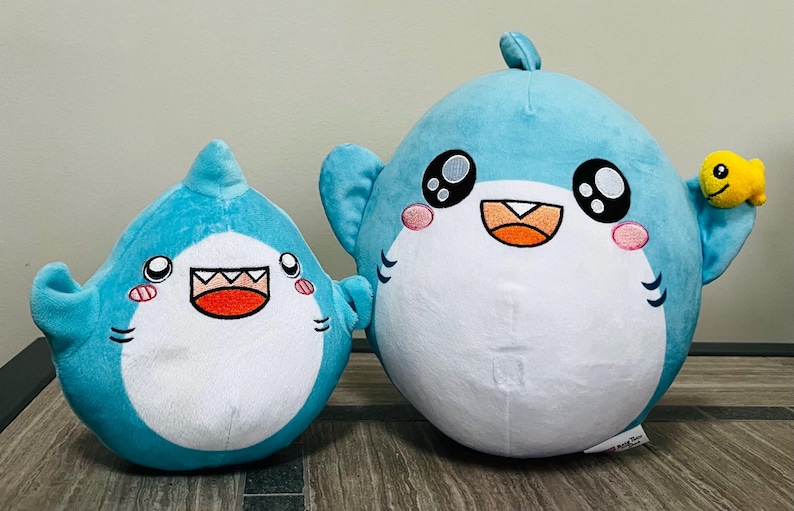 May include: Two plush shark toys in shades of blue and white. The sharks have large, expressive eyes, pink cheeks, and open mouths. One shark has a small yellow fish attached. The toys are designed with a cartoonish aesthetic.
